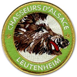 Ecusson rond brodé du club de chasseurs d'Alsace. Il représente une tête de sanglier de profil, gueule ouverte, entouré d'un bandeau vert tout autour, sur lequel il est écrit Chasseurs d'Alsace, Leutenheim en blanc . la bordure de l'écusson est couleur or.