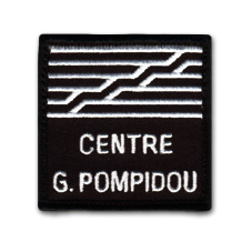 Ecusson brodé du centre Georges Pompidou. Il est carré, avec un fond noir sur lequel est brodé en blanc le texte Centre G. pompidou en lettres capitales en bas. Au dessus, des lignes horizontales blanches et deux lignes formant un escalier rappellent la façade de ce centre d'art contemporain.